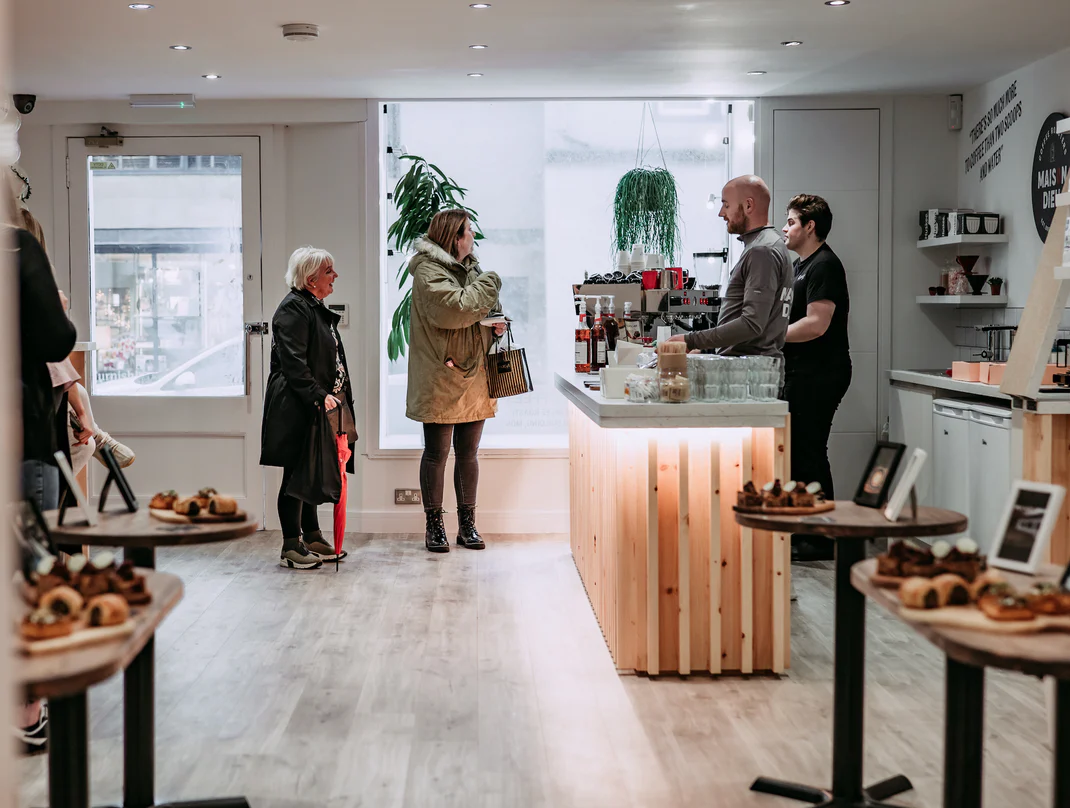 Interior view of a coffee shop with customers interacting at the counter and pastries displayed on tables.